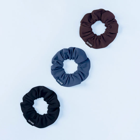 3-pack fabric scrunchies: dark brown, teal, and black with stitched DRYNE labels